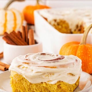 Closeup of a pumpkin cinnamon roll on a plate topped with cream cheese frosting, with cinnamon sticks and small pumpkins in the background