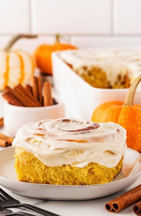 Closeup of a pumpkin cinnamon roll on a plate topped with cream cheese frosting, with cinnamon sticks and small pumpkins in the background