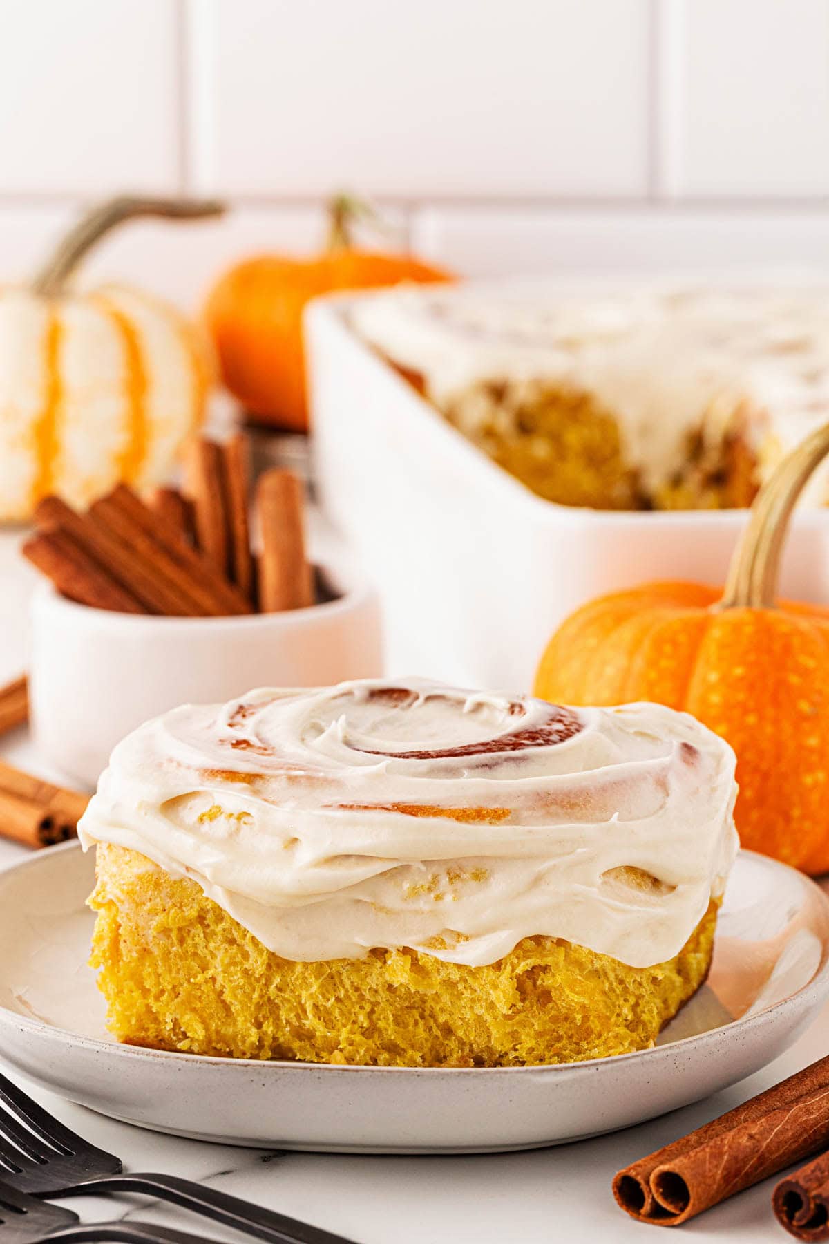 Closeup of a pumpkin cinnamon roll on a plate topped with cream cheese frosting, with cinnamon sticks and small pumpkins in the background
