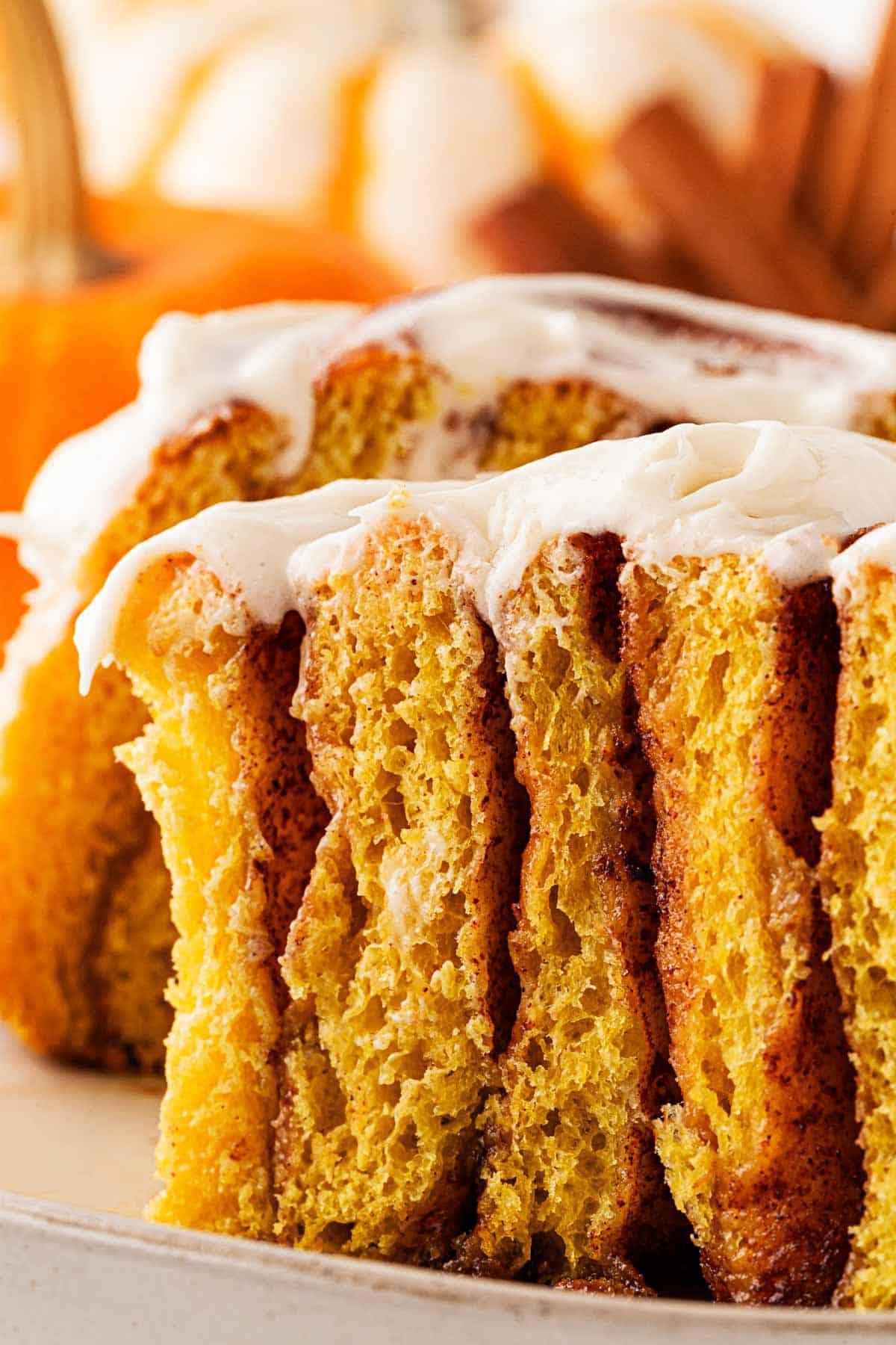 A sliced pumpkin cinnamon roll on a plate, showing its soft texture and cream cheese frosting, with pumpkins and cinnamon sticks in the background