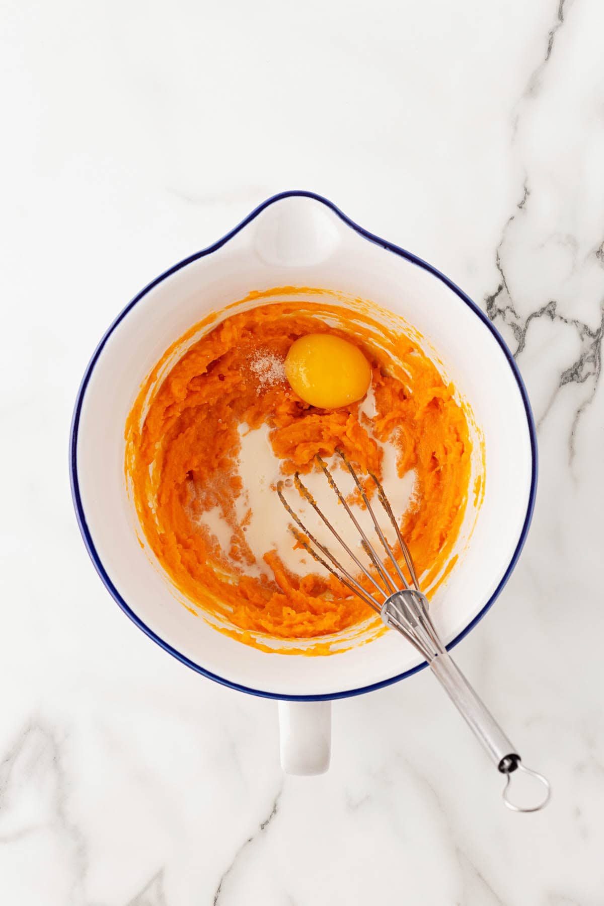 Mashed sweet potato in a mixing bowl with an egg, milk, and a whisk ready to blend