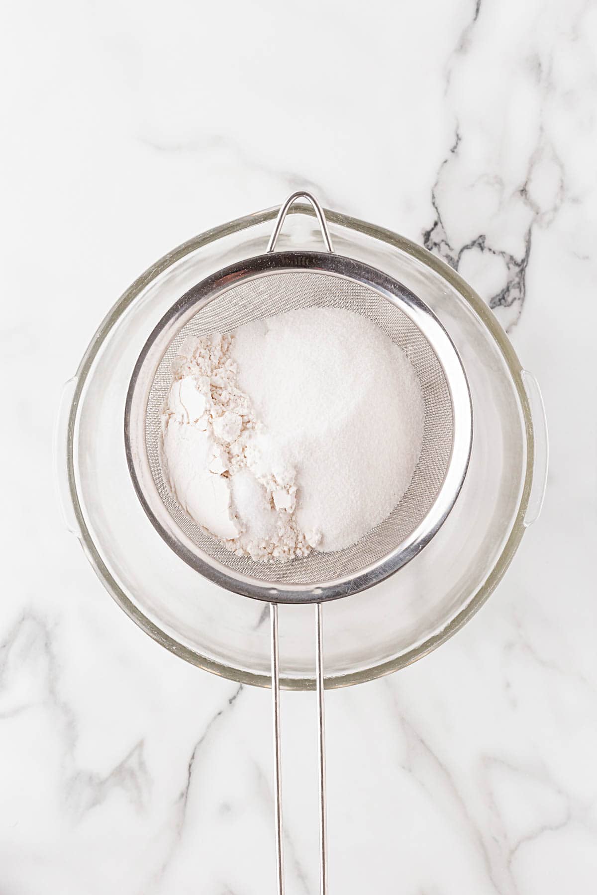 Sugar, salt and flour being sifted together into a glass mixing bowl with a fine mesh sieve