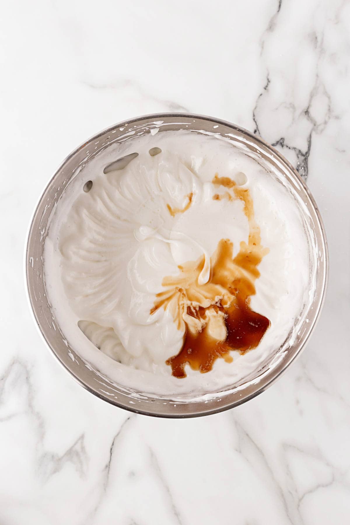 Whipped egg whites in a metal mixing bowl with extracts added on top