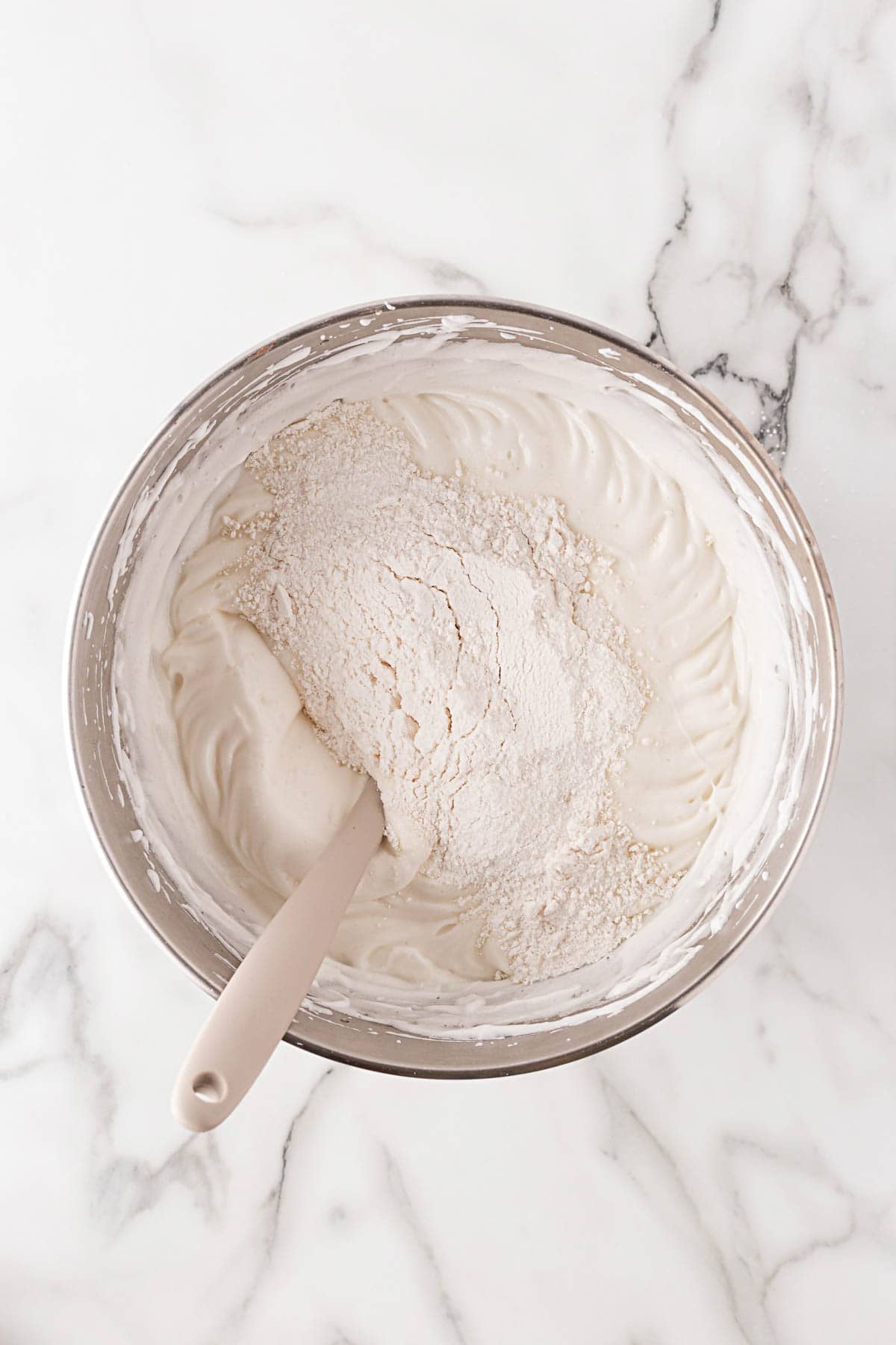 Batter being folded with flour into whipped egg whites using a spatula in a metal mixing bowl