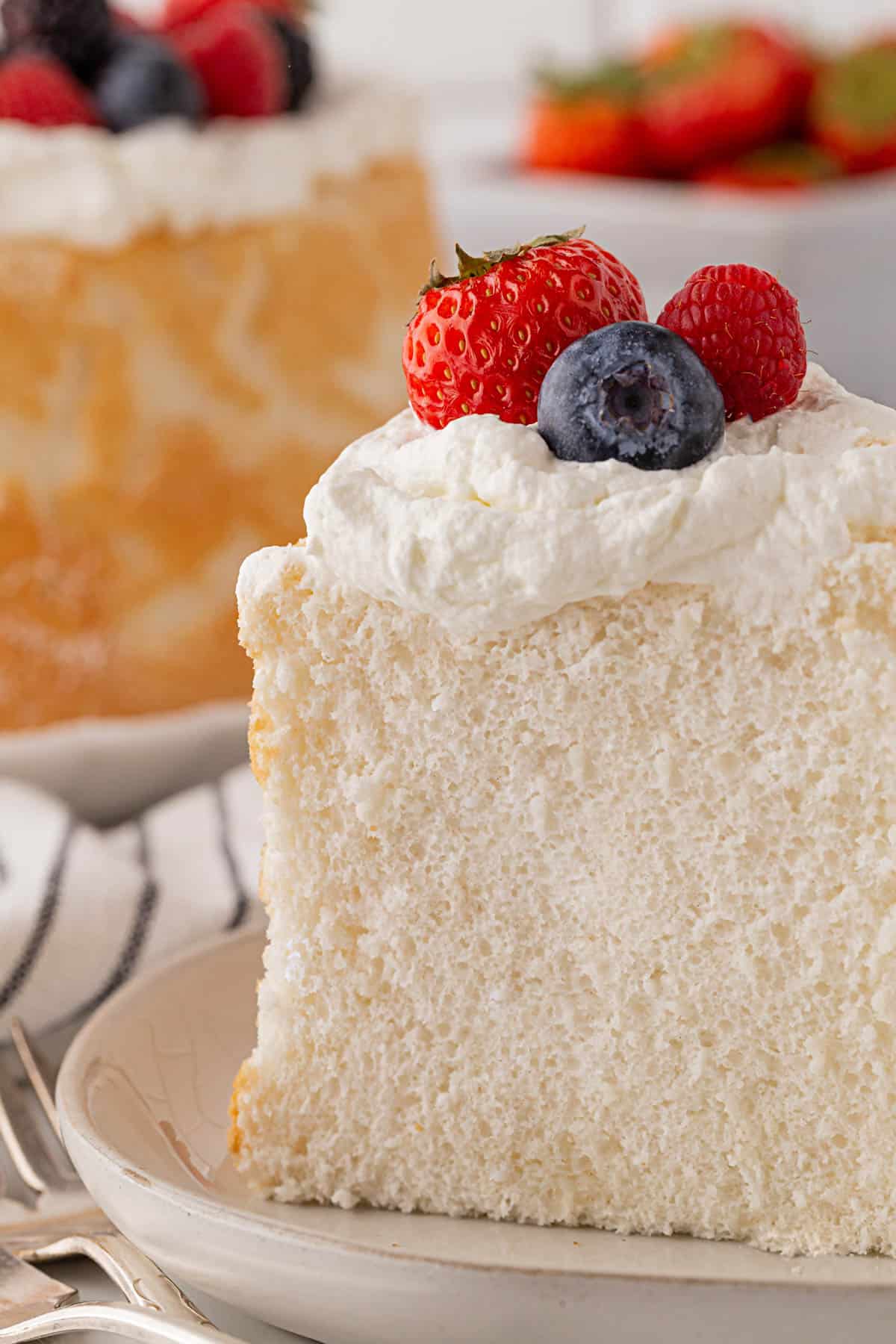 Closeup of a single slice of angel food cake on a plate topped with whipped cream and fresh berries, showing the tender texture of the cake