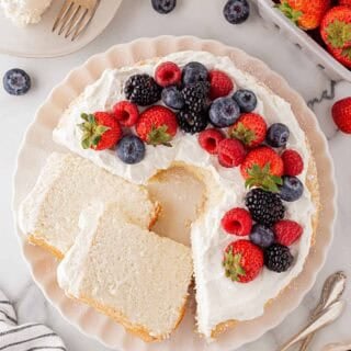 Decorated angel food cake topped with whipped cream and fresh berries including strawberries, raspberries, blackberries, and blueberries, with slices cut and served on plates