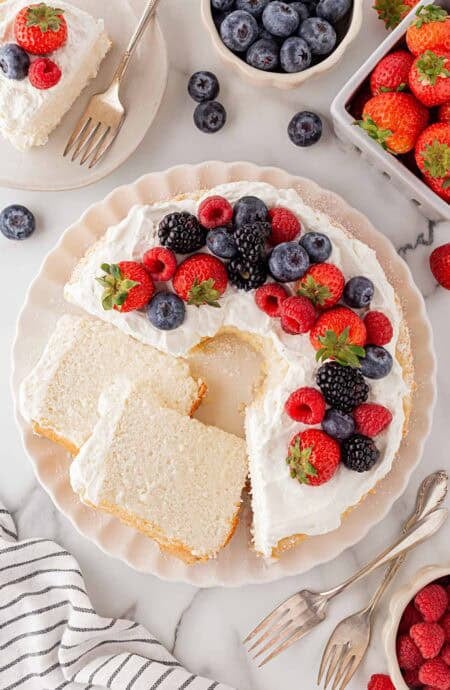 Decorated angel food cake topped with whipped cream and fresh berries including strawberries, raspberries, blackberries, and blueberries, with slices cut and served on plates