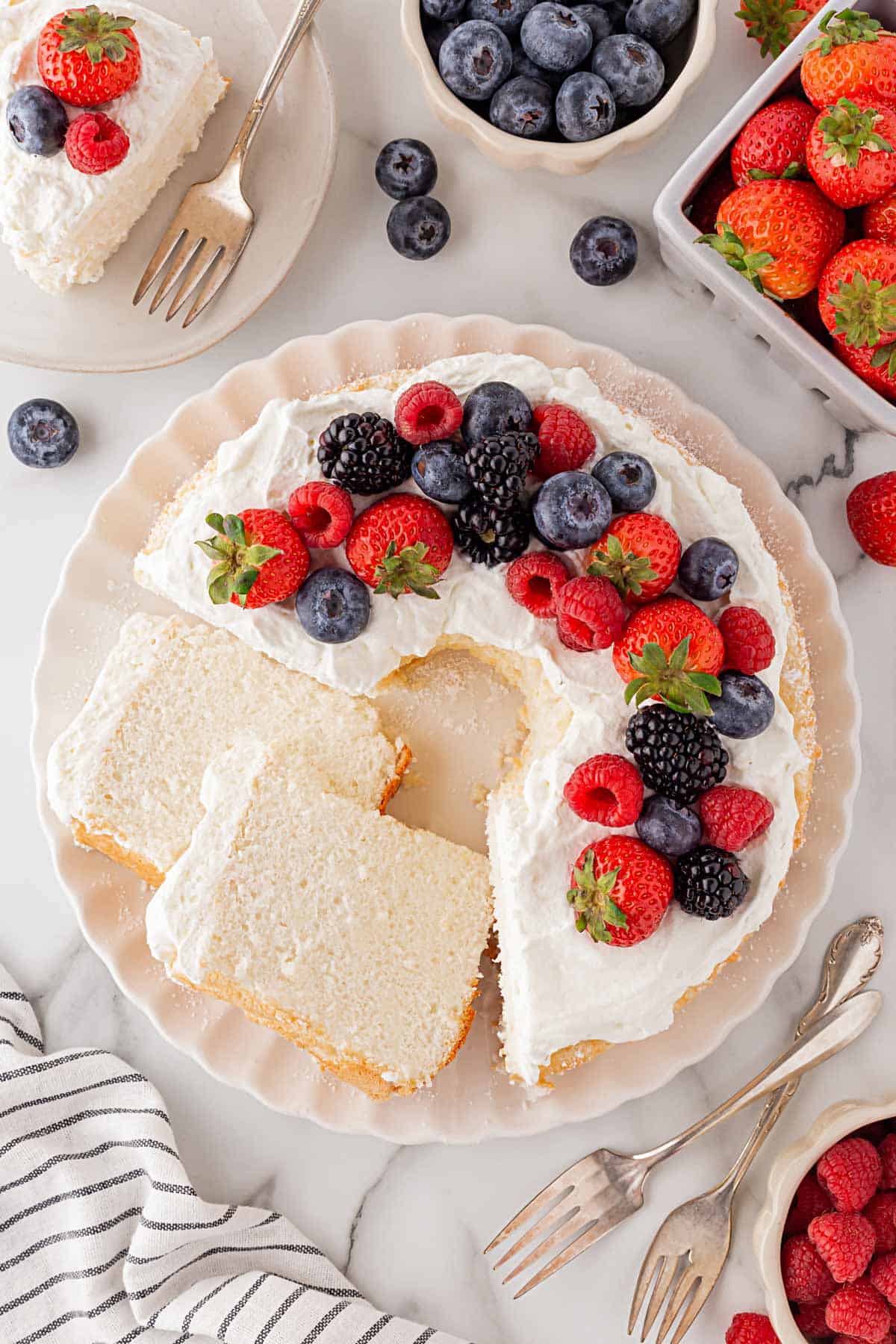 Decorated angel food cake topped with whipped cream and fresh berries including strawberries, raspberries, blackberries, and blueberries, with slices cut and served on plates