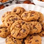 A white scalloped plate piled with golden brown butter pecan cookies with chocolate chips and pecans