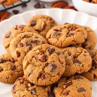 A white scalloped plate piled with golden brown butter pecan cookies with chocolate chips and pecans