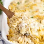 A wooden spoon lifting a creamy serving of chicken alfredo casserole with rotini pasta from a white baking dish