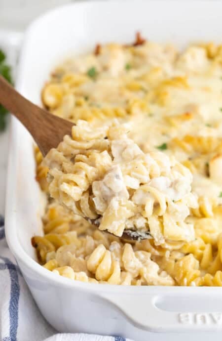 A wooden spoon lifting a creamy serving of chicken alfredo casserole with rotini pasta from a white baking dish