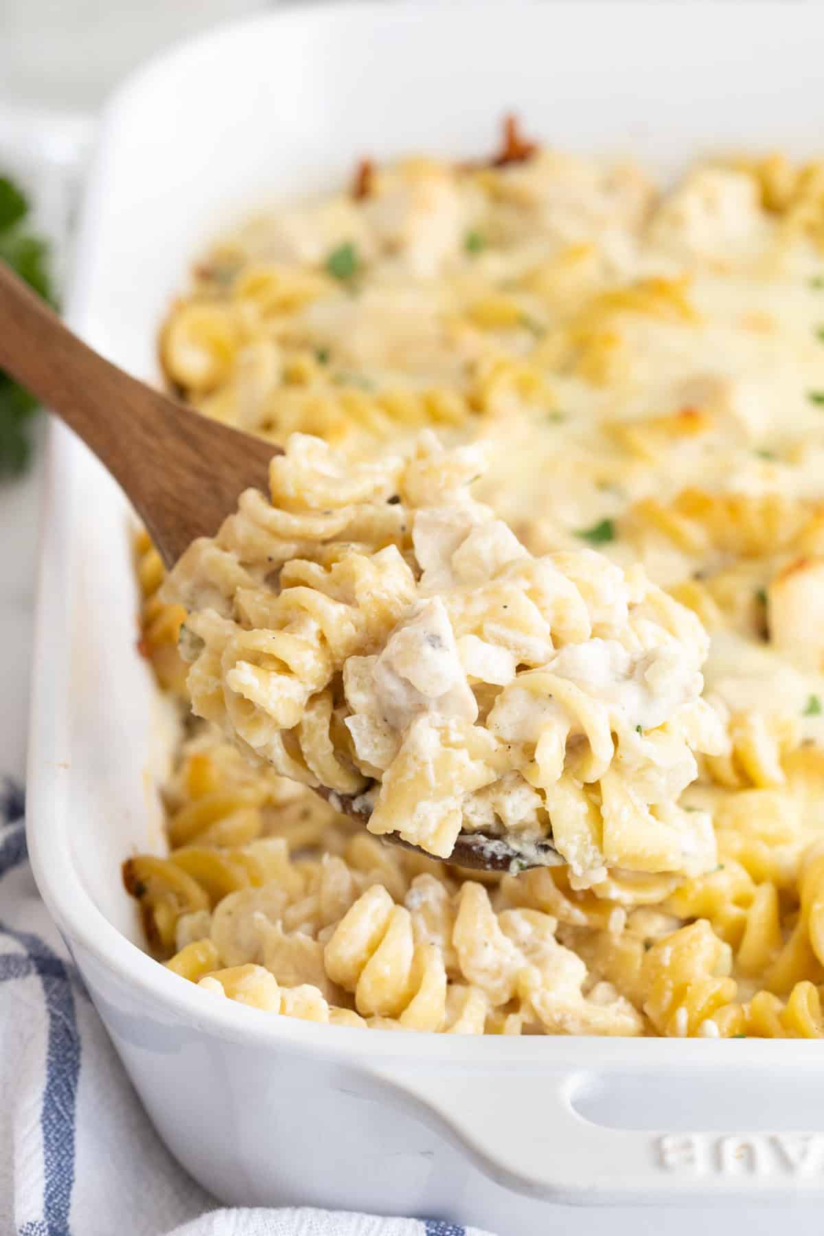 A wooden spoon lifting a creamy serving of chicken alfredo casserole with rotini pasta from a white baking dish
