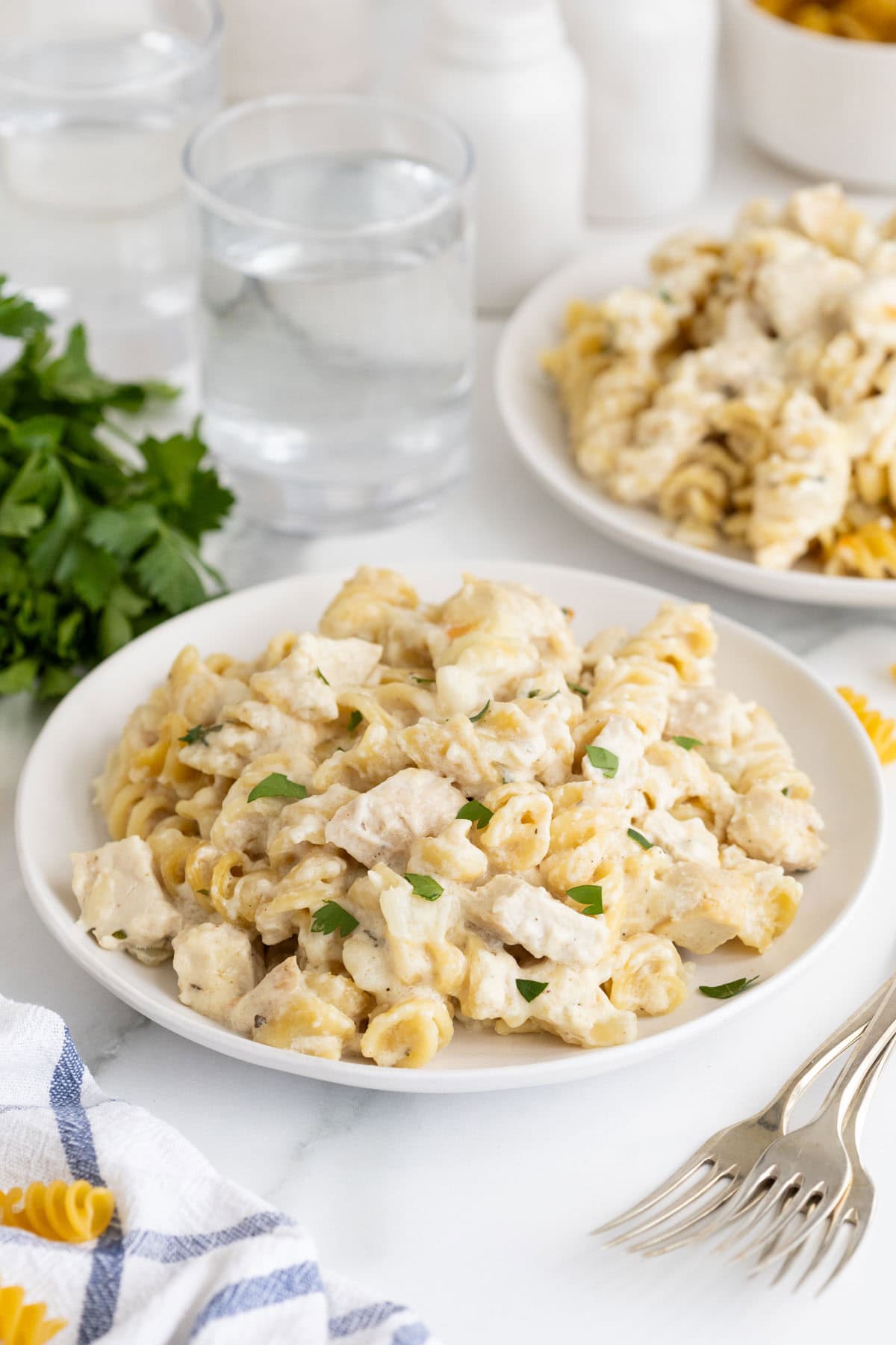 A plate of creamy chicken alfredo casserole garnished with fresh parsley, with a second plate and glasses of water in the background