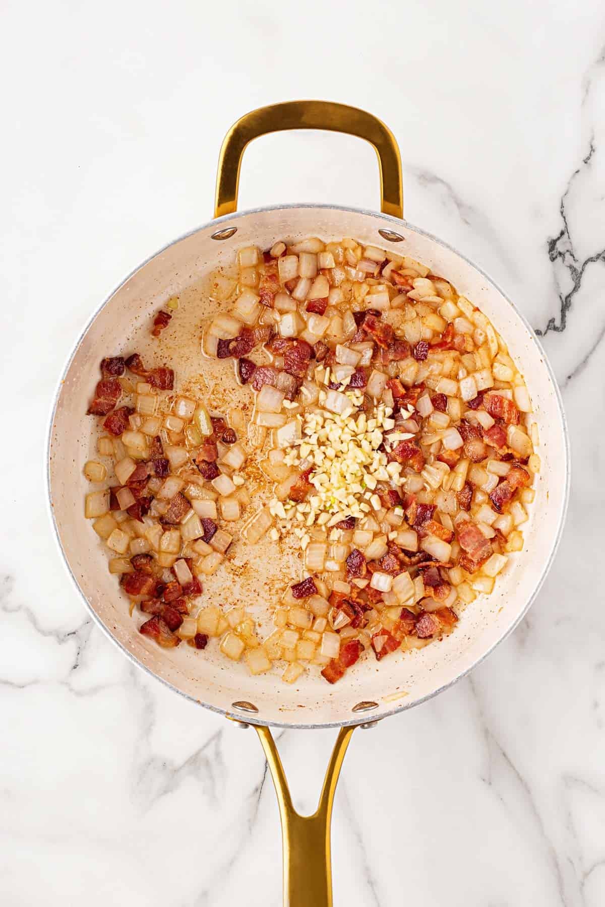Skillet filled with sautéed onions, bacon pieces, and minced garlic on a marble countertop