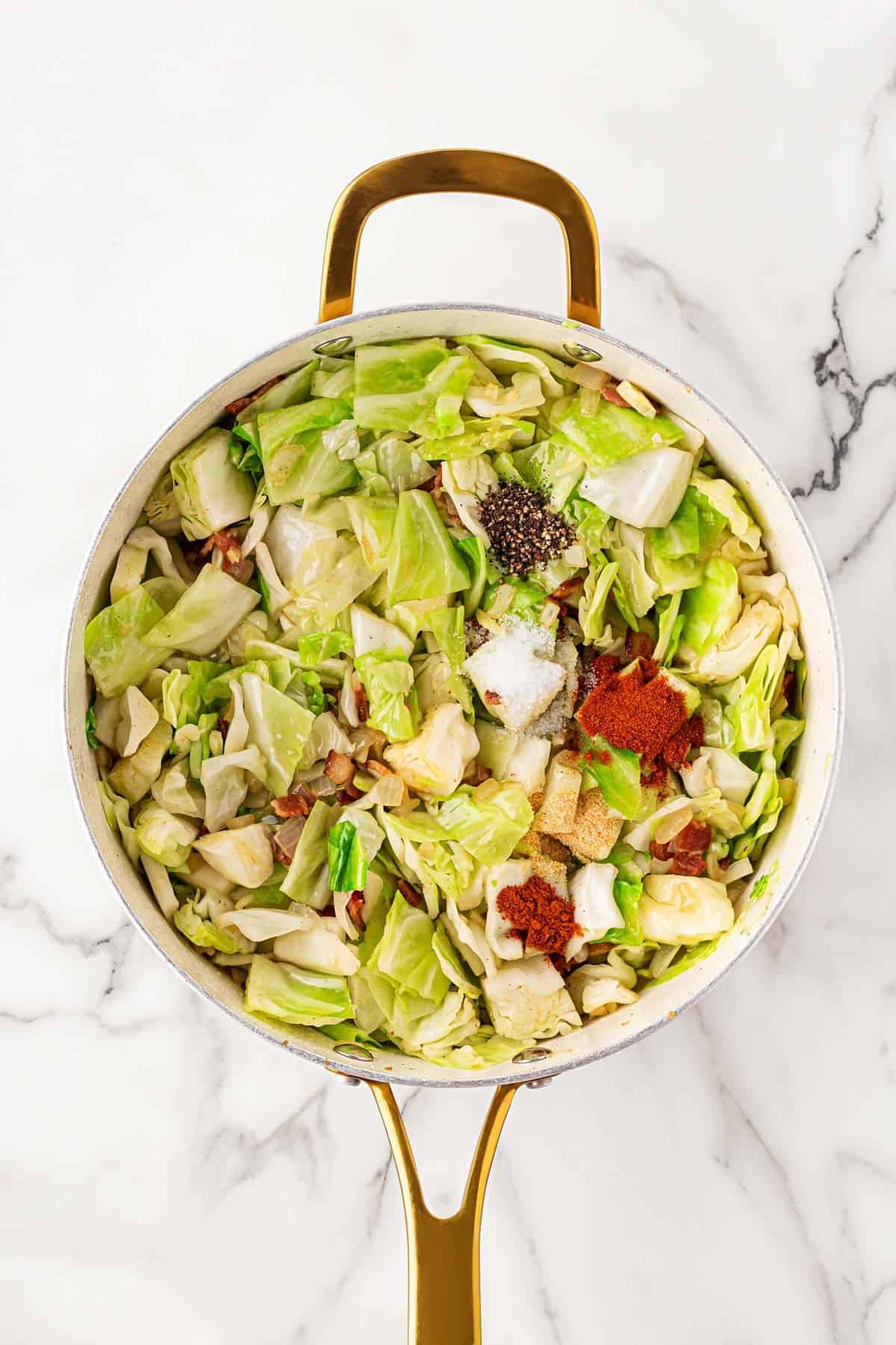 Chopped cabbage in a skillet with bacon, onions, and a mix of spices including paprika, salt, and pepper