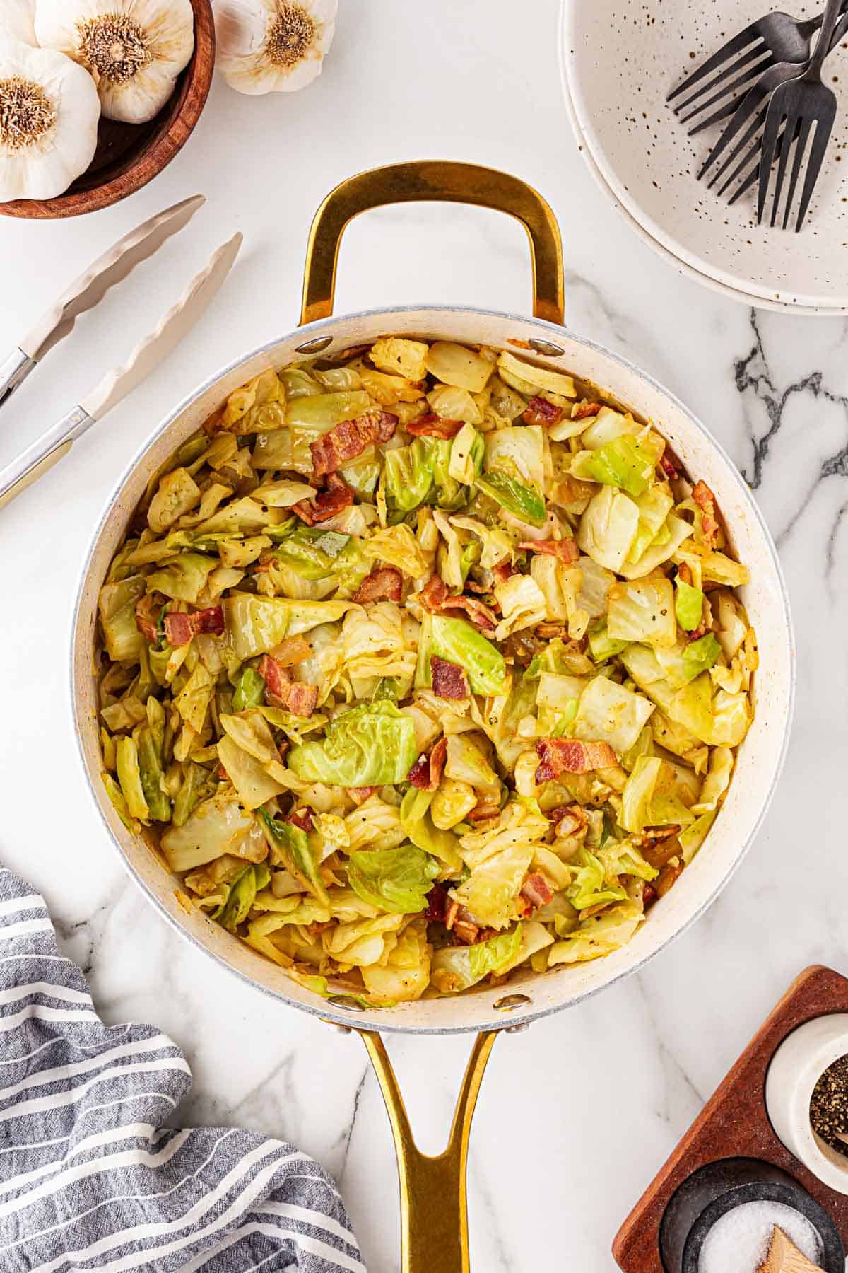 Skillet of golden fried cabbage with bacon, resting on a marble surface beside garlic bulbs and utensils