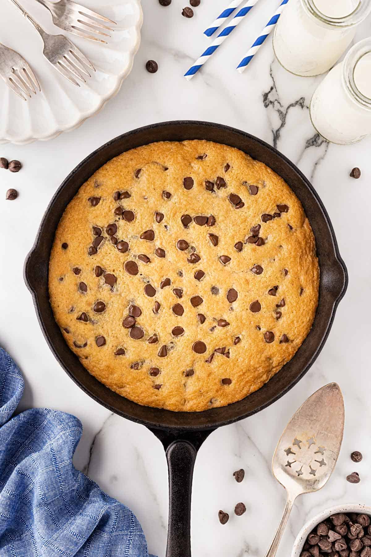 Baked chocolate chip skillet cookie golden brown on top, surrounded by milk bottles, forks, and a blue napkin
