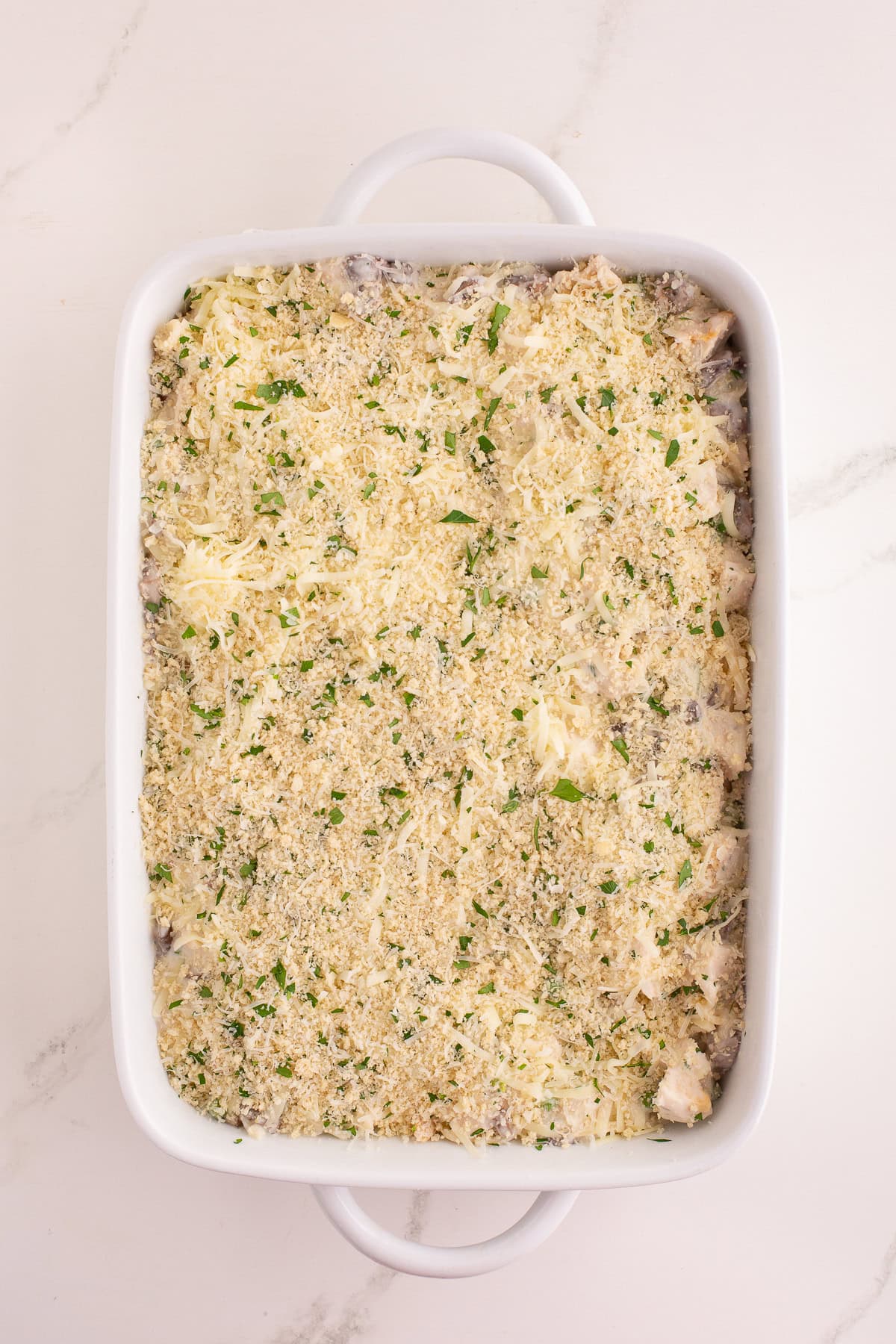 Unbaked turkey tetrazzini mixture spread in a casserole dish, topped with breadcrumbs, cheese, and herbs