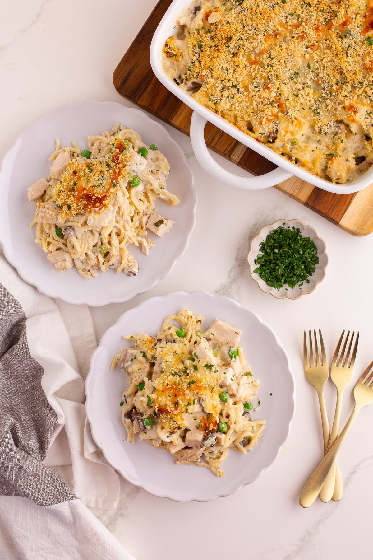 Two plates of creamy turkey tetrazzini served beside the casserole dish, garnished with parsley and golden breadcrumbs