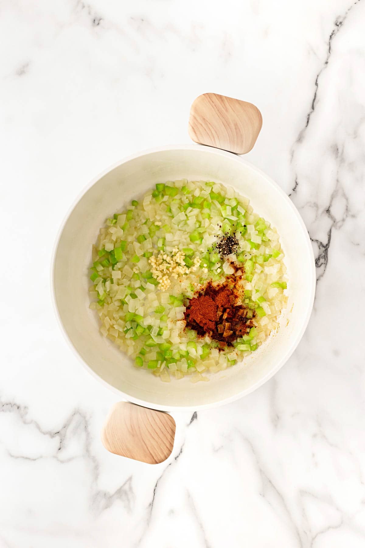 Chopped onions, celery, garlic, and spices cooking in a white pot on a marble surface