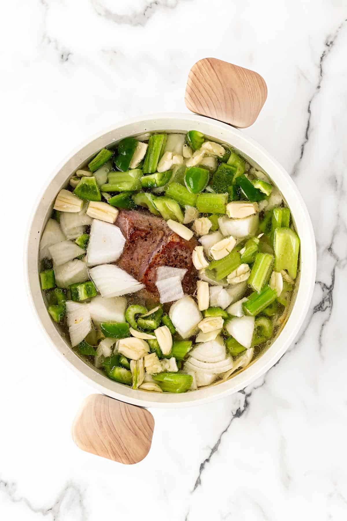 Pork shoulder simmering in broth with chopped onions, celery, and garlic