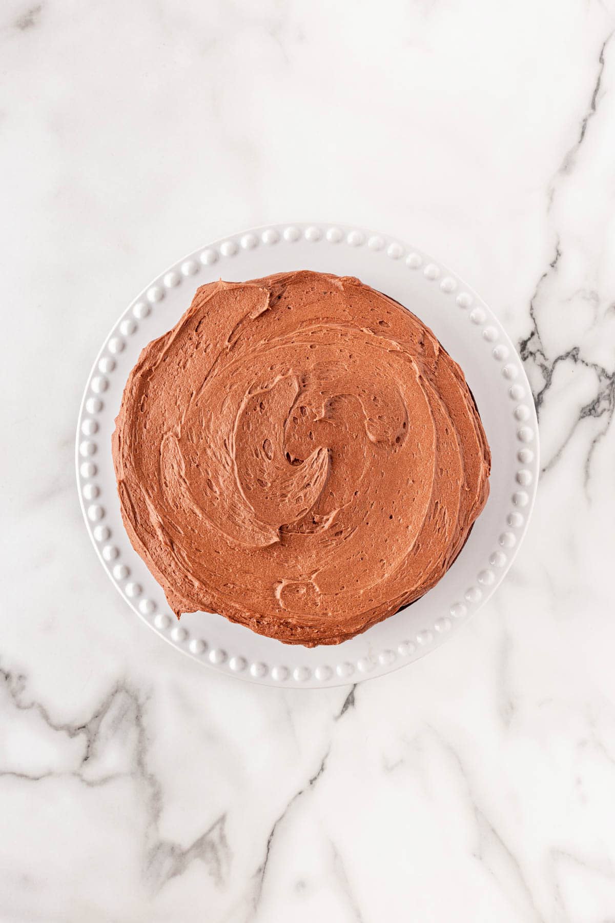 Chocolate cake layer fully frosted with smooth chocolate buttercream on a white cake stand
