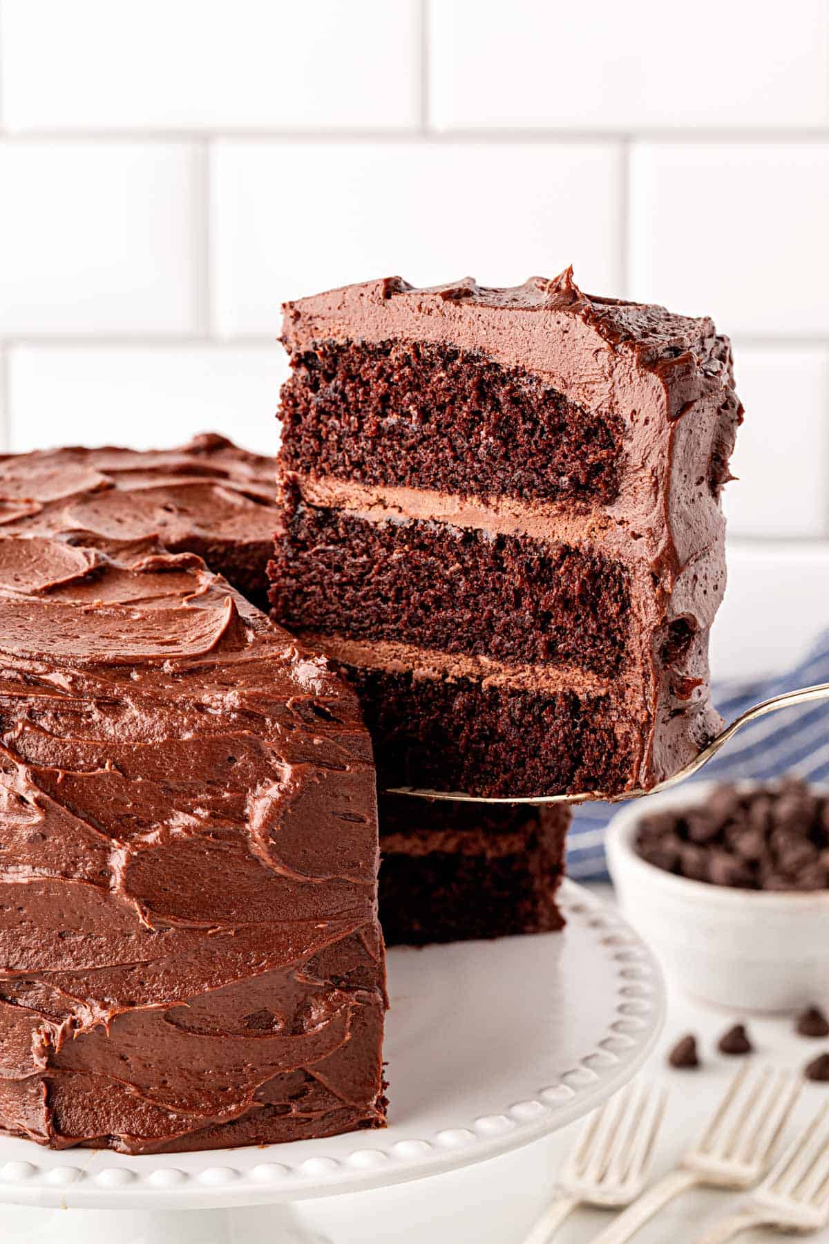 Slice of triple layer chocolate cake being lifted from a frosted cake on a white cake stand