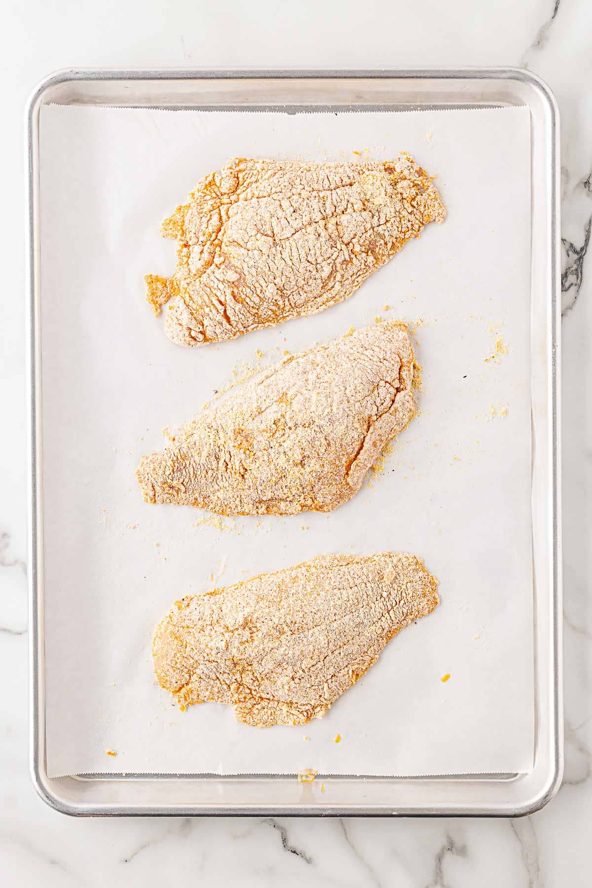 Three fish fillets coated in seasoned flour on a parchment-lined baking sheet