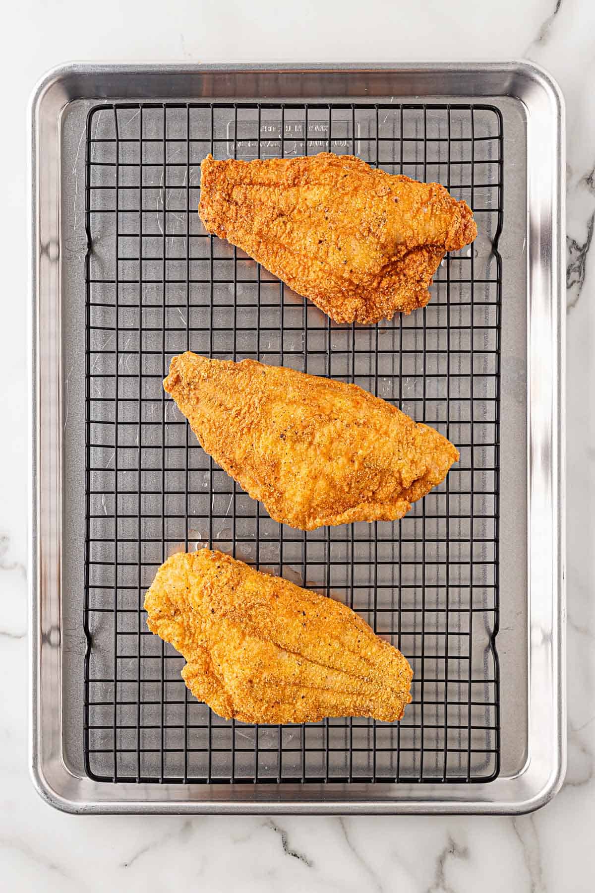 Three breaded fish fillets resting on a wire rack over a sheet pan