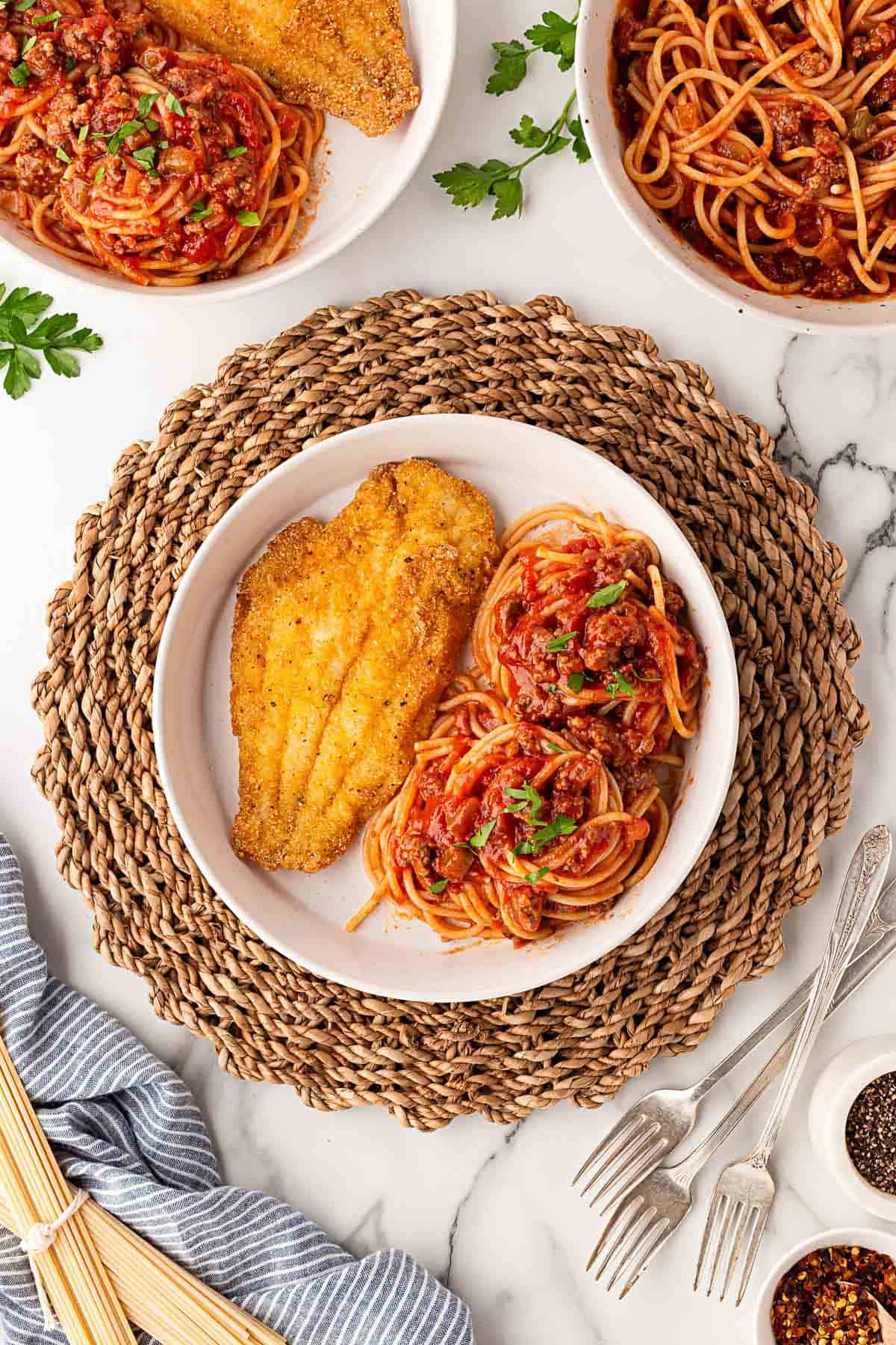 Breaded fried fish fillet served with spaghetti and meat sauce on a plate, with more plates of fish and spaghetti next to it