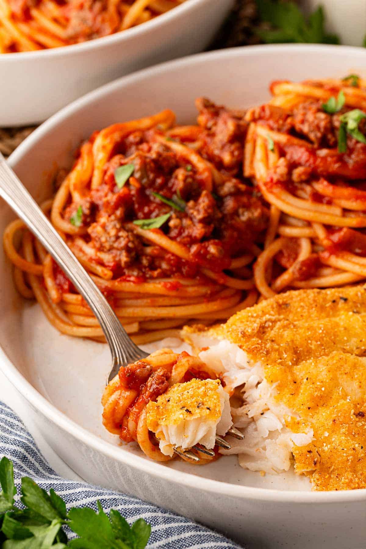 Fork holding a bite of spaghetti and fried fish from a plate of fish and spaghetti