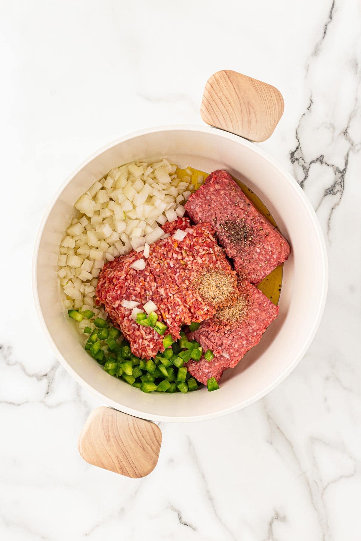 Bowl with ground beef, diced onions, green peppers, oil, and seasonings ready to cook