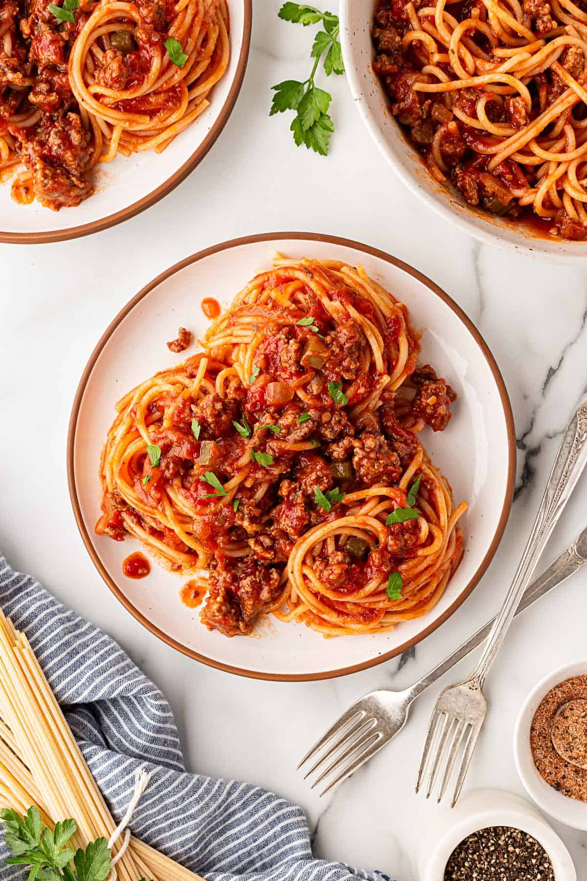 Overhead shot of a plate of spaghetti with homemade meat sauce and parsley, surrounded by bowls of spaghetti and uncooked pasta