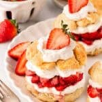 Strawberry shortcakes layered with whipped cream and strawberries on a plate, with more strawberries, shortcakes, and biscuits in the background