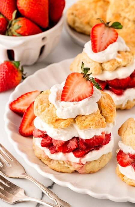 Strawberry shortcakes layered with whipped cream and strawberries on a plate, with more strawberries, shortcakes, and biscuits in the background