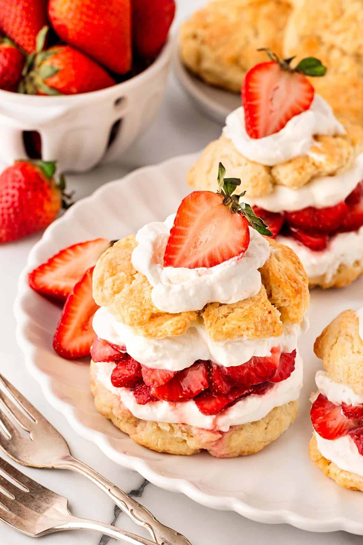 Strawberry shortcakes layered with whipped cream and strawberries on a plate, with more strawberries, shortcakes, and biscuits in the background