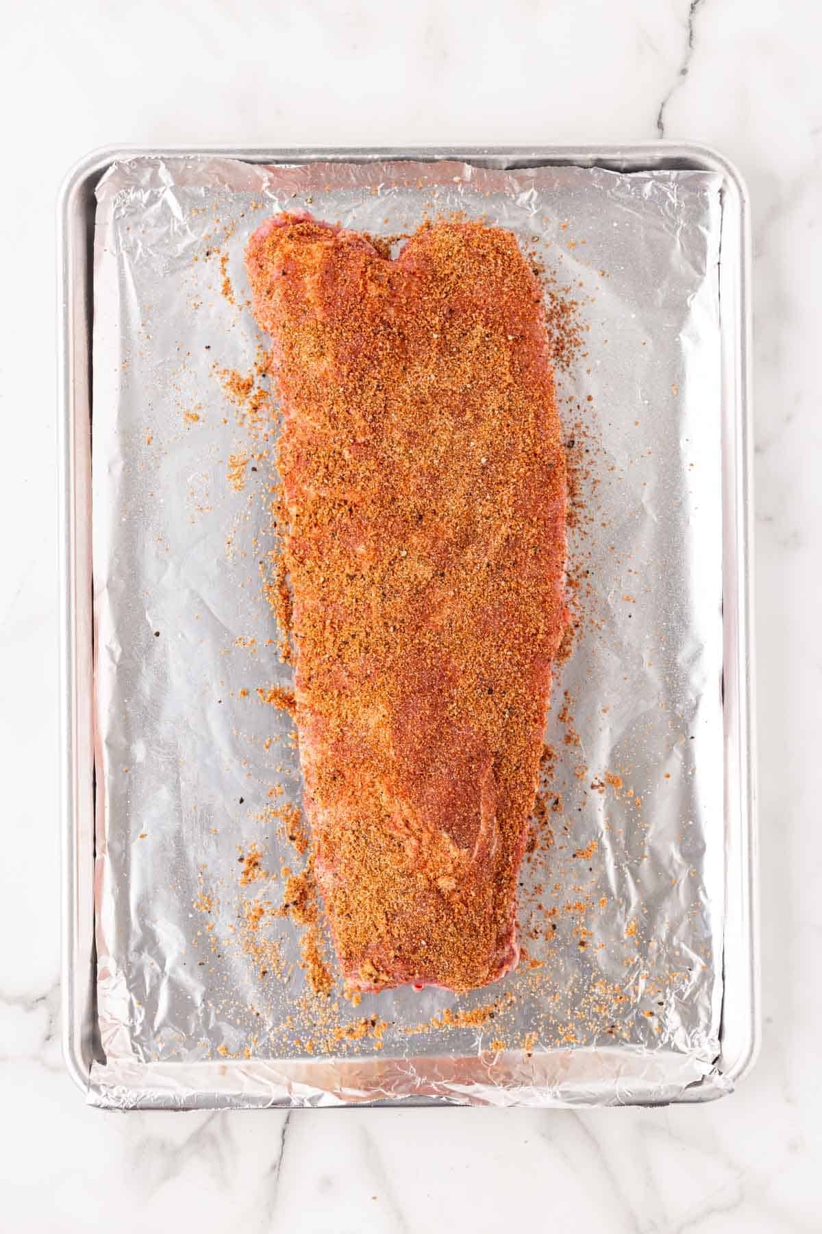 Rack of pork ribs coated in dry rub on a foil-lined baking sheet