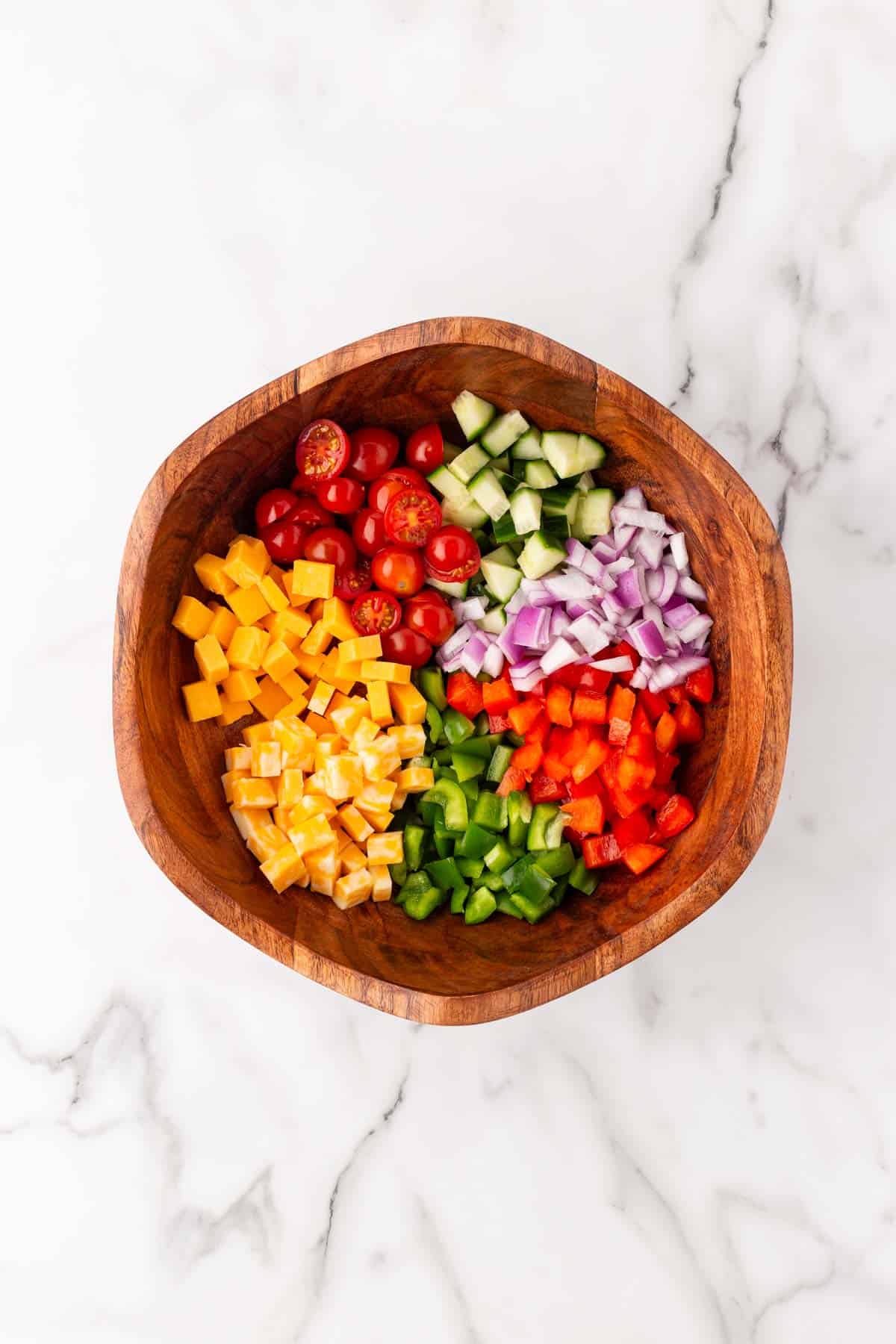 Chopped cucumbers, cherry tomatoes, red onion, bell peppers, and cheese cubes arranged in a wooden bowl