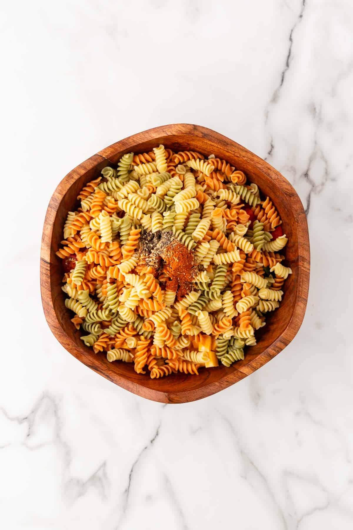Tricolor rotini pasta in a wooden bowl topped with seasonings before mixing