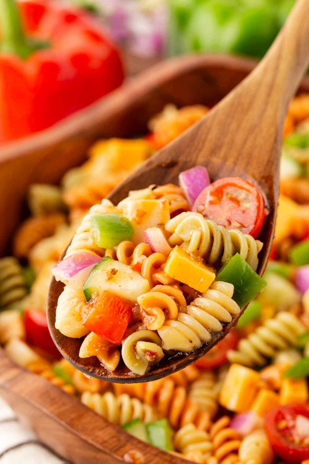 Wooden spoon scooping colorful pasta salad with rotini, cheese cubes, cucumbers, tomatoes, and bell peppers