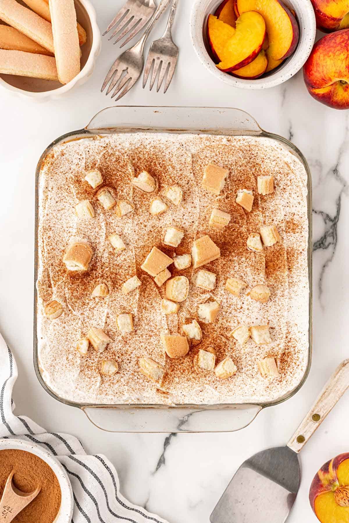 Overhead shot of peach cobbler tiramisu topped with cinnamon and ladyfingers, with peaches, ladyfingers, and spices surrounding it
