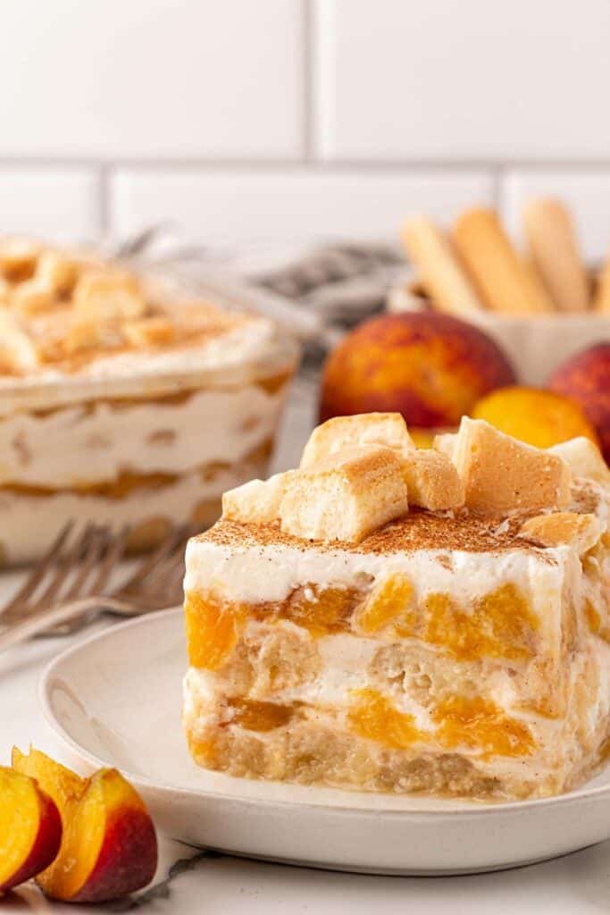 Peach cobbler tiramisu with creamy layers, peaches, and ladyfingers, with more tiramisu and peaches in the background