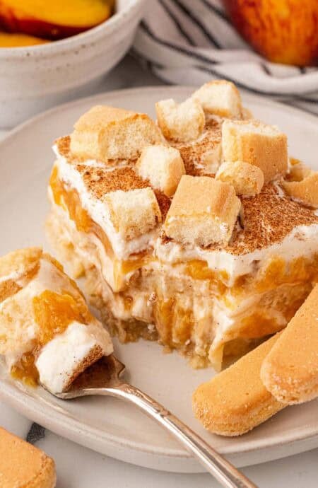 Fork holding a creamy bite of peach cobbler tiramisu beside a layered slice topped with cinnamon and ladyfingers