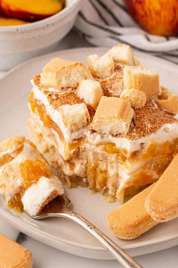 Fork holding a creamy bite of peach cobbler tiramisu beside a layered slice topped with cinnamon and ladyfingers