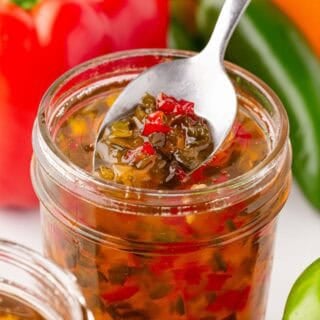 Spoon scooping pepper jelly from an open mason jar with peppers in the background
