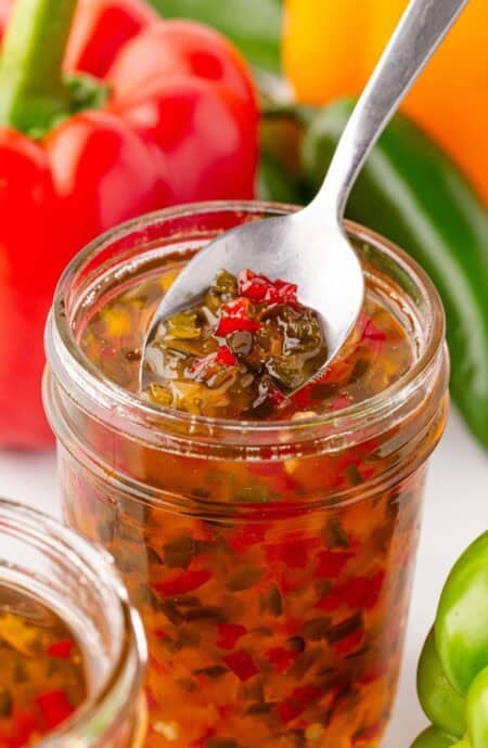 Spoon scooping pepper jelly from an open mason jar with peppers in the background