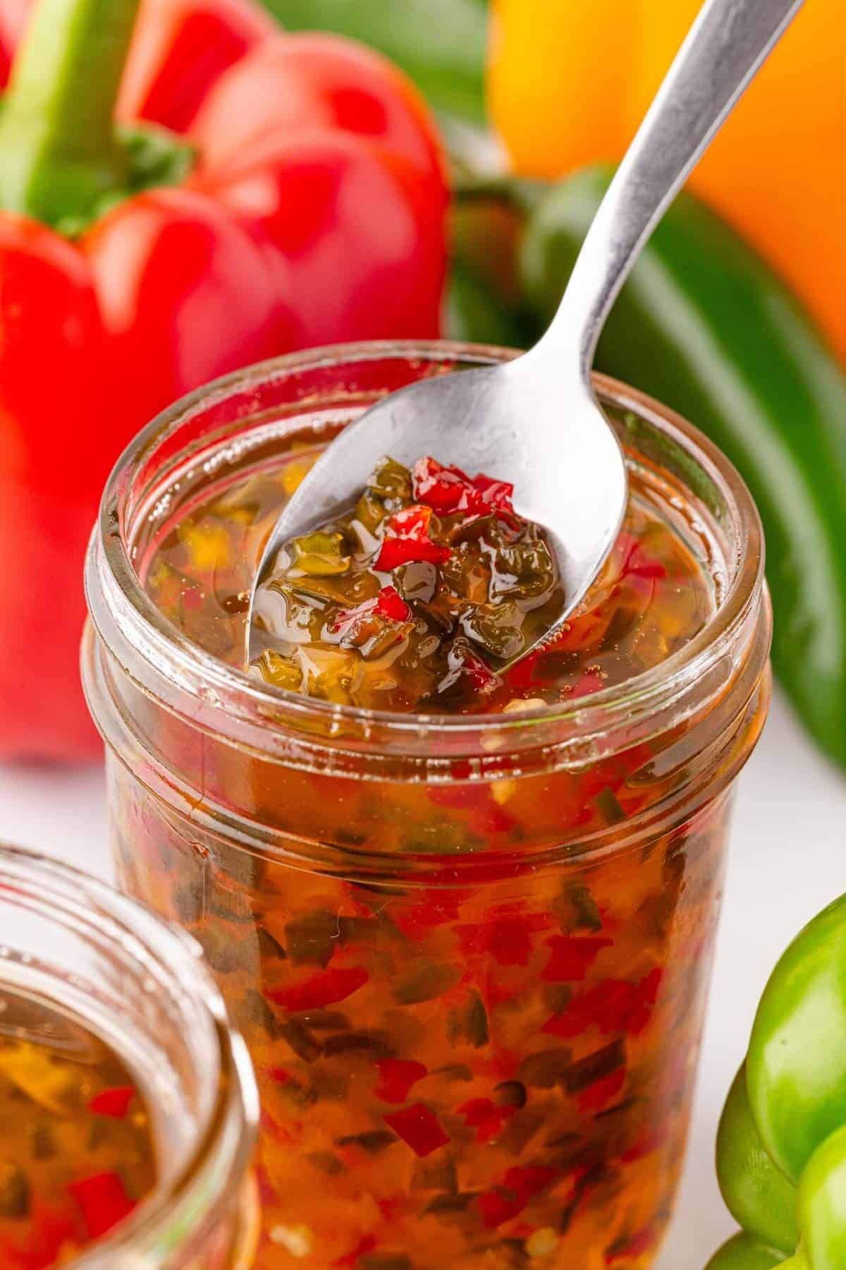 Spoon scooping pepper jelly from an open mason jar with peppers in the background