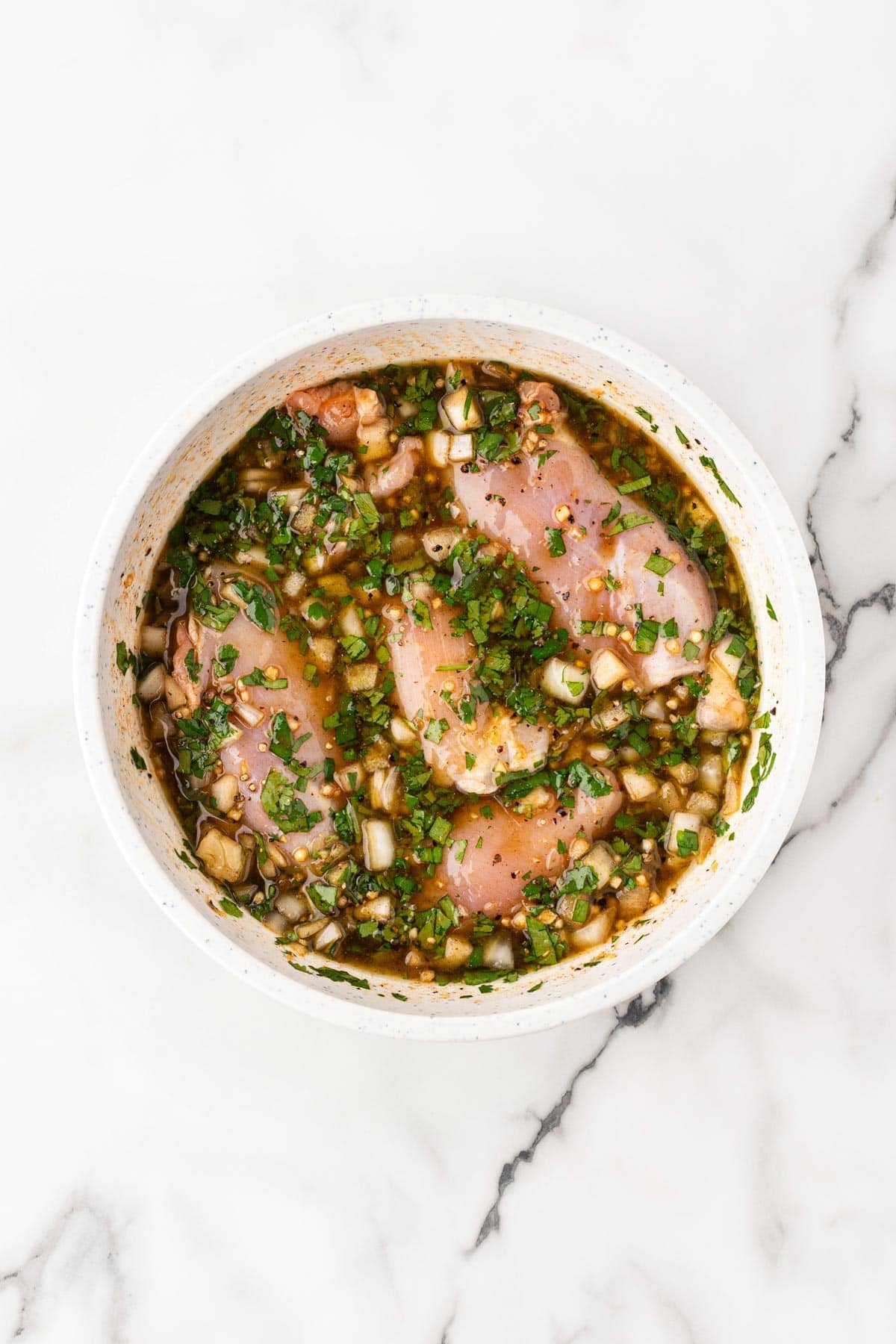 Raw chicken thighs marinating in a herb-filled sauce inside a white bowl on a white marble background