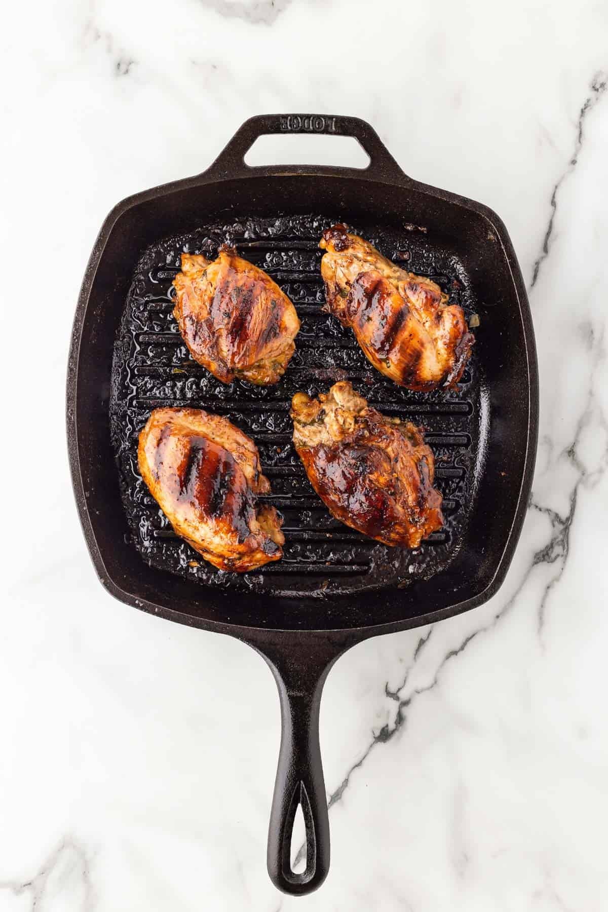 Grilled chicken thighs with char marks in a cast iron grill pan on a white marble background